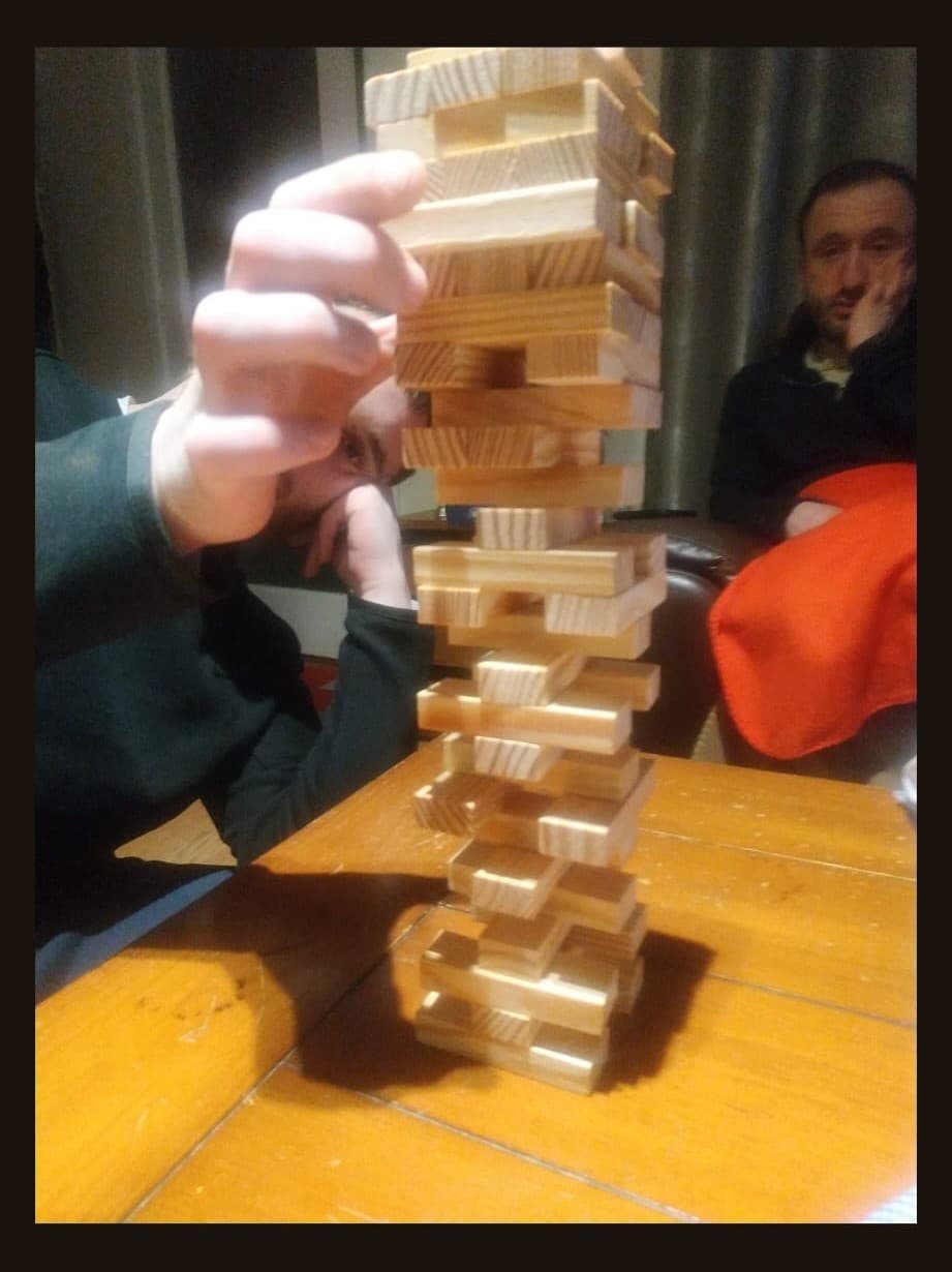 A person leaning beside a tall wooden block tower while another person sits nearby.